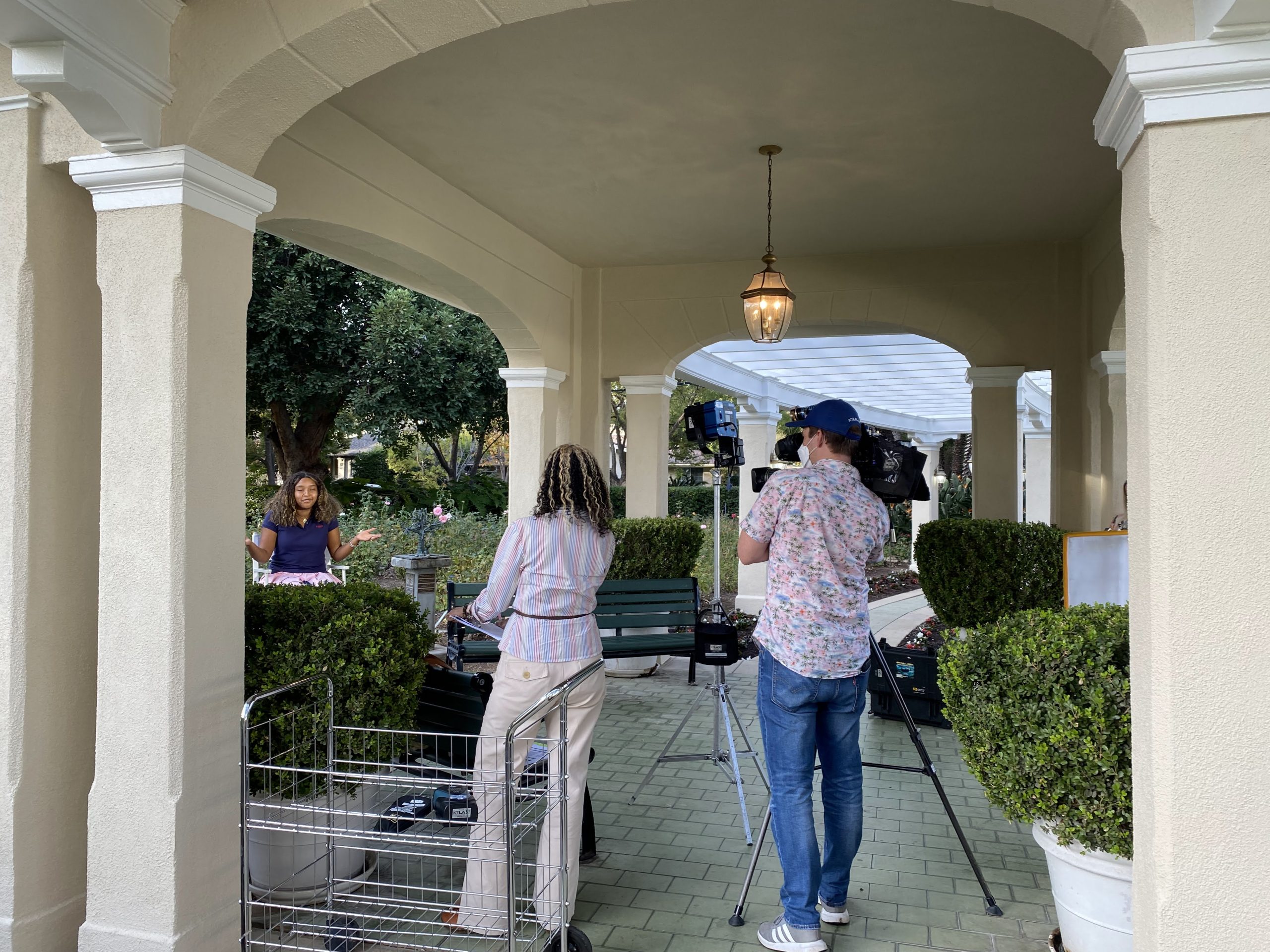 KTLA camera operator films 2022 Rose Princess McKenzie Street at Tournament House during an outdoor interview. Rear shot of field producer Patricia Nazario, photographer and lighting equipment positioned beneath a covered garden walkway.