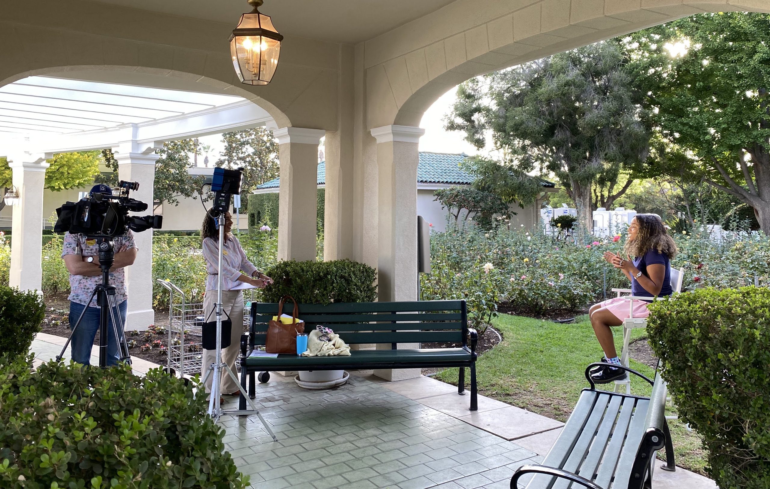 KTLA camera operator films 2022 Rose Princess McKenzie Street at Tournament House during an outdoor interview. Field Producer Patricia Nazario and lighting equipment positioned beneath a covered garden walkway.