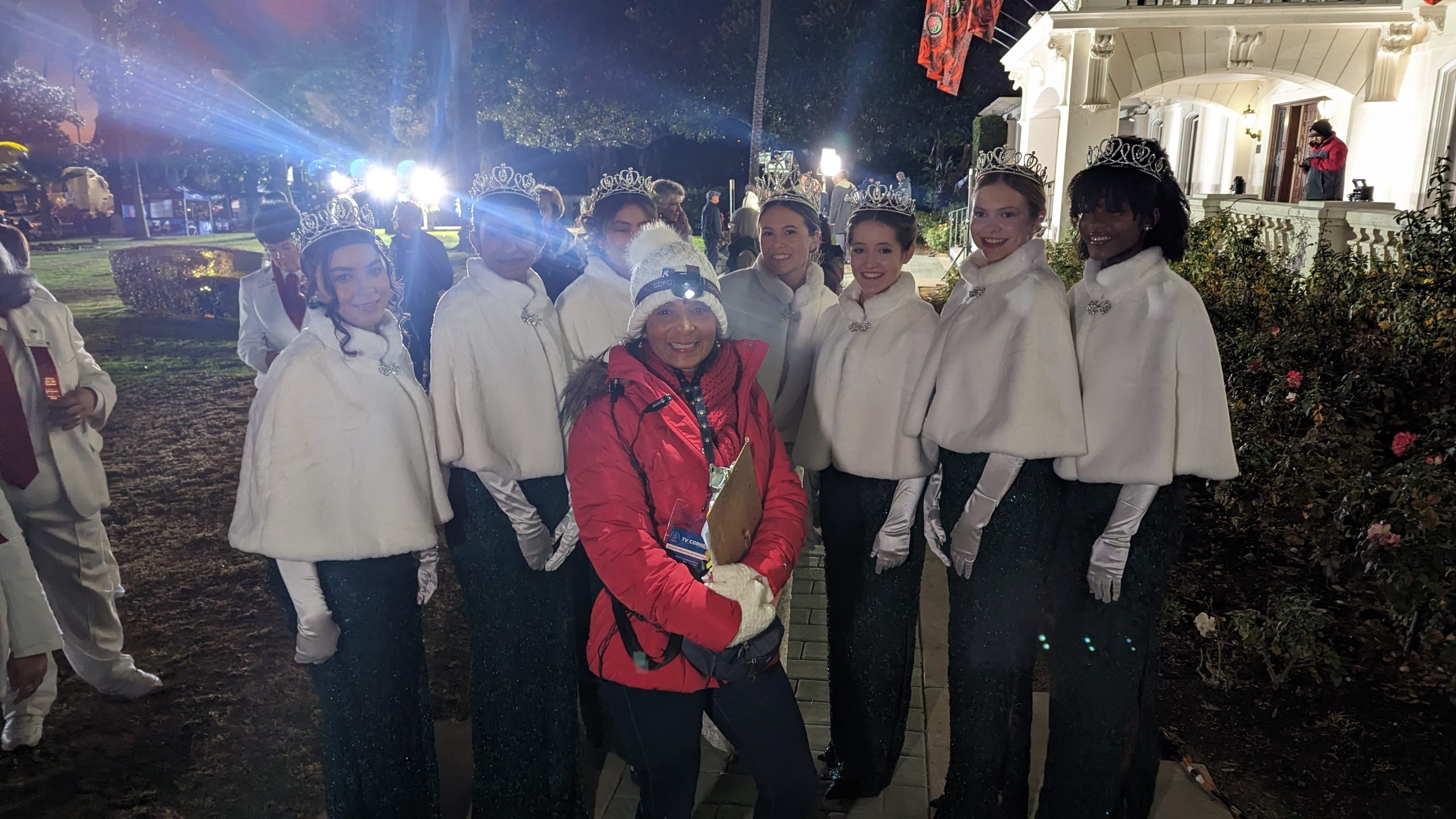 KTLA field producer Patricia Nazario poses with the 2023 Rose Queen and Royal Court, wearing tiaras and white capes, during predawn Rose Parade preparations in Pasadena.