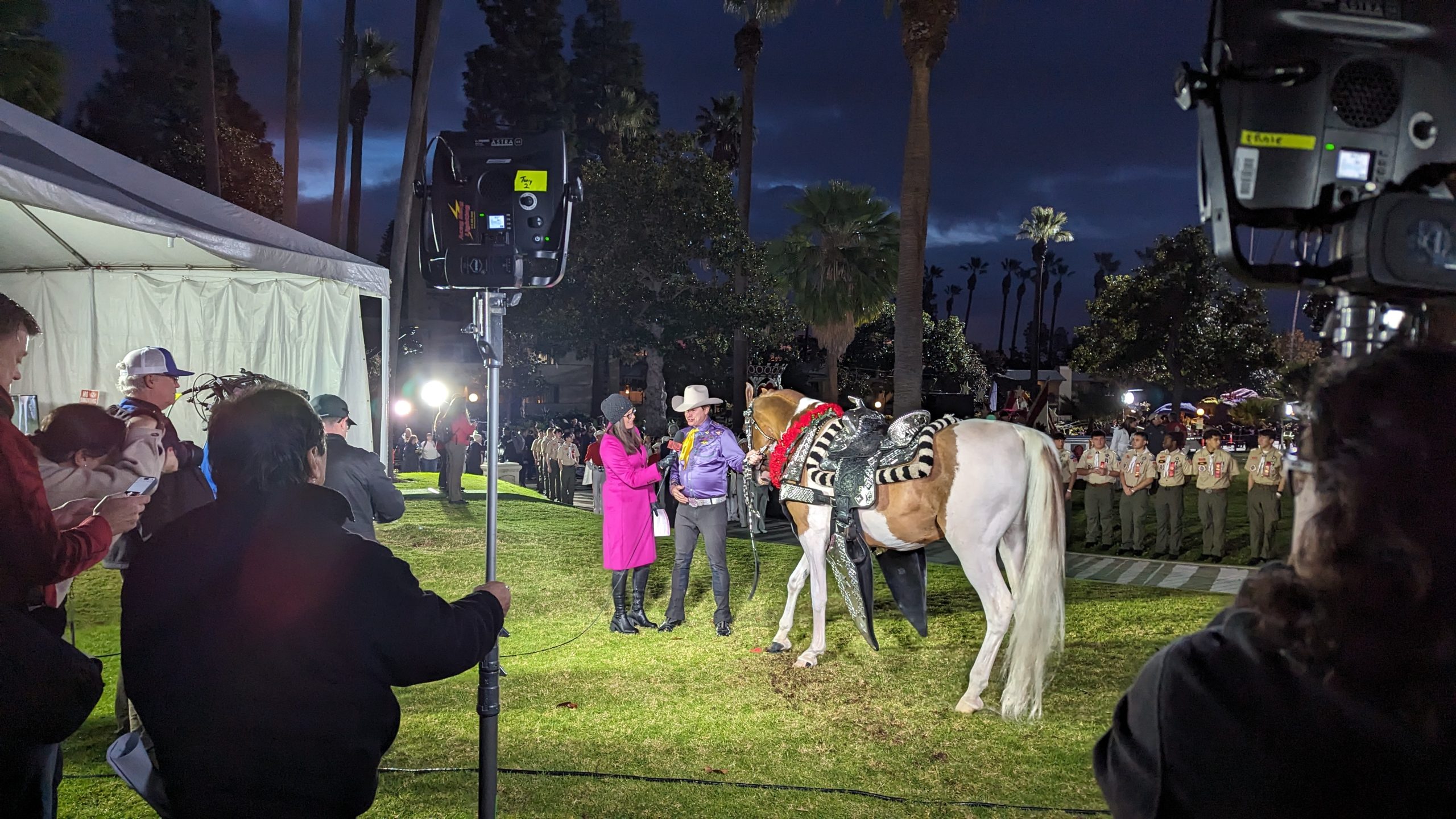 KTLA television reporter in a bright pink coat interviewing a cowboy beside a decorated horse at night, surrounded by broadcast cameras, lighting equipment and production crew in Pasadena.
