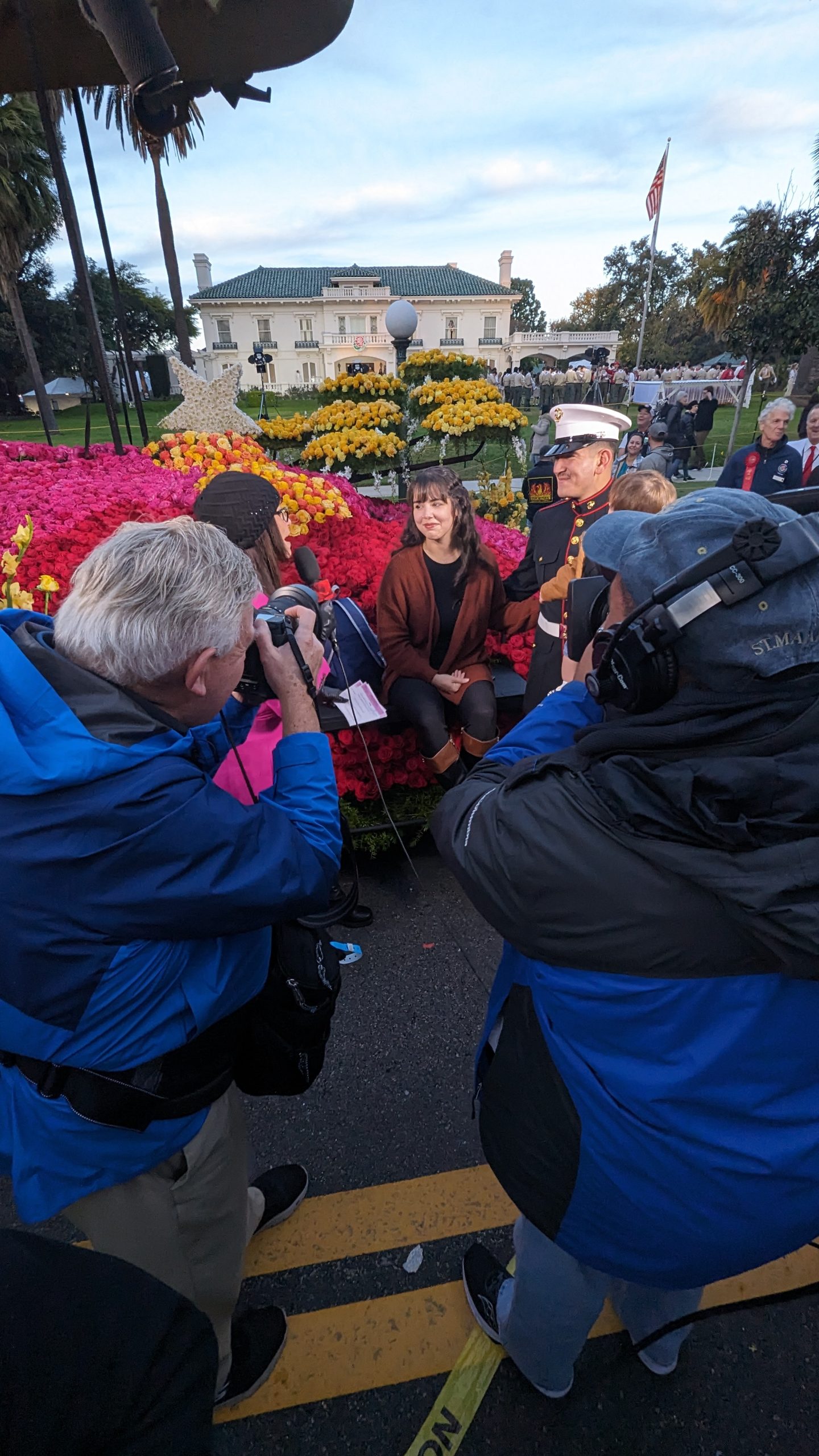 KTLA television crew filming an interview with a U.S. Marine in dress uniform and his wife, seated beside a colorful floral display outside Tournament House in Pasadena during the 2024 Rose Parade festivities.
