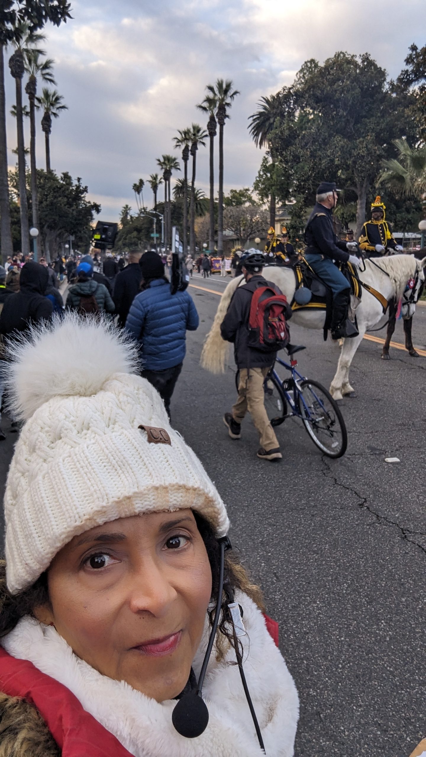 KTLA field producer Patricia Nazario wearing a white knit hat, red jacket and communications headset on the 2024 Rose Parade route, with mounted equestrian participants and production personnel behind her.