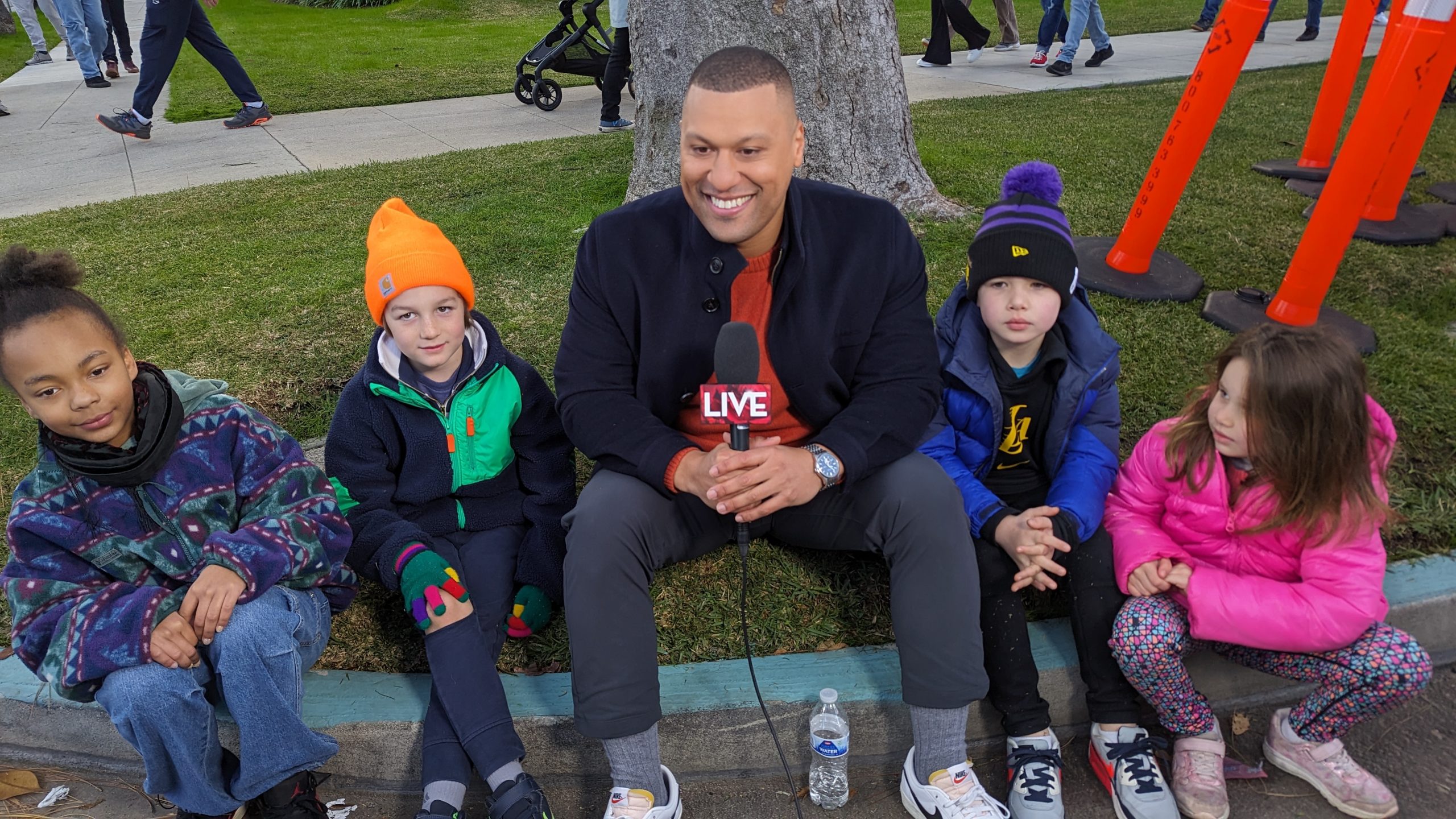 KTLA's Pedro Rivera seated on a curb with four children dressed in winter jackets, holding a microphone marked LIVE during the 2024 Rose Parade festivities in Pasadena.