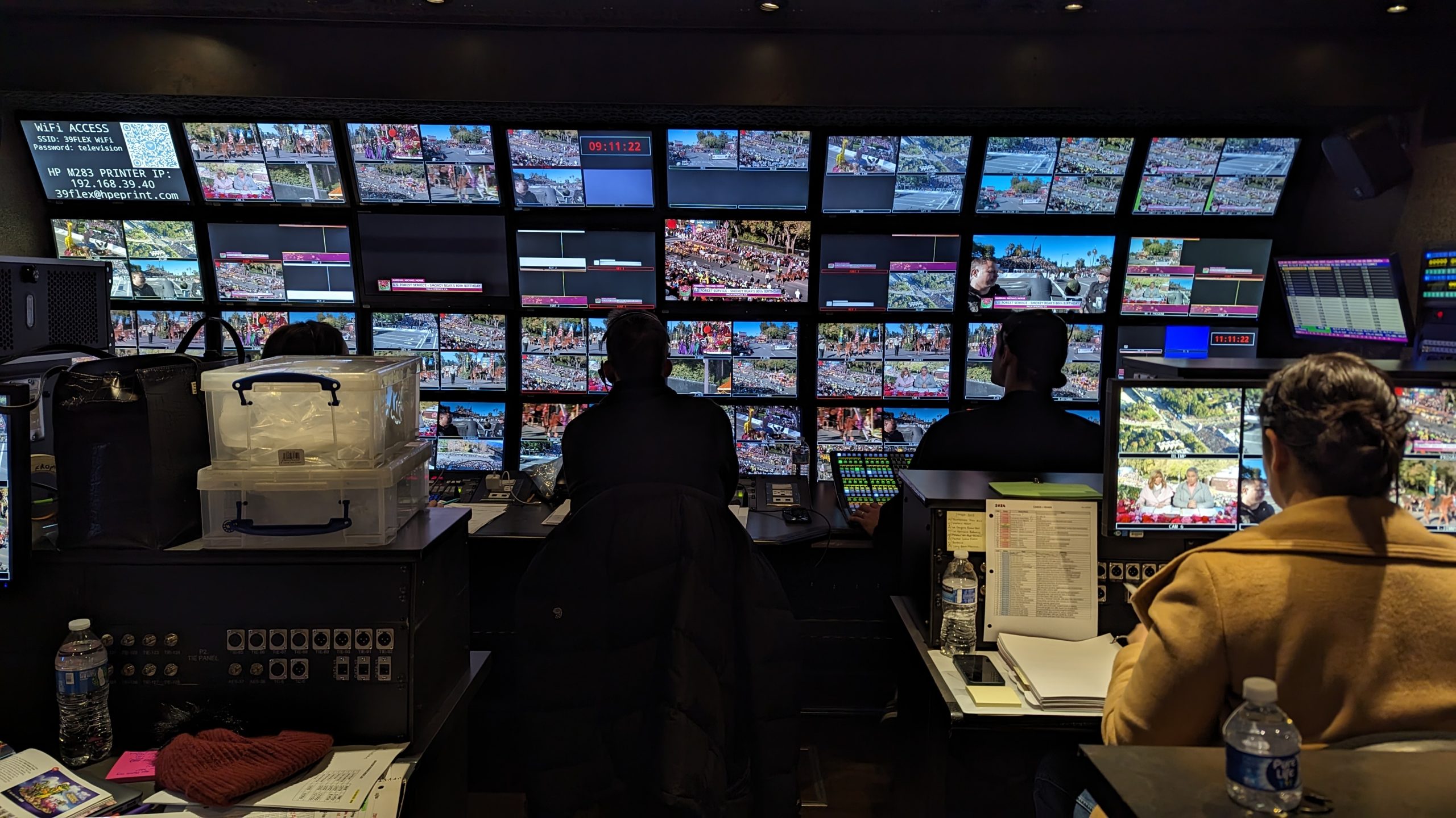 Wide view inside KTLA's 2024 Rose Parade broadcast control room, showing production personnel working at consoles beneath a wall of television monitors displaying live parade footage and graphics.