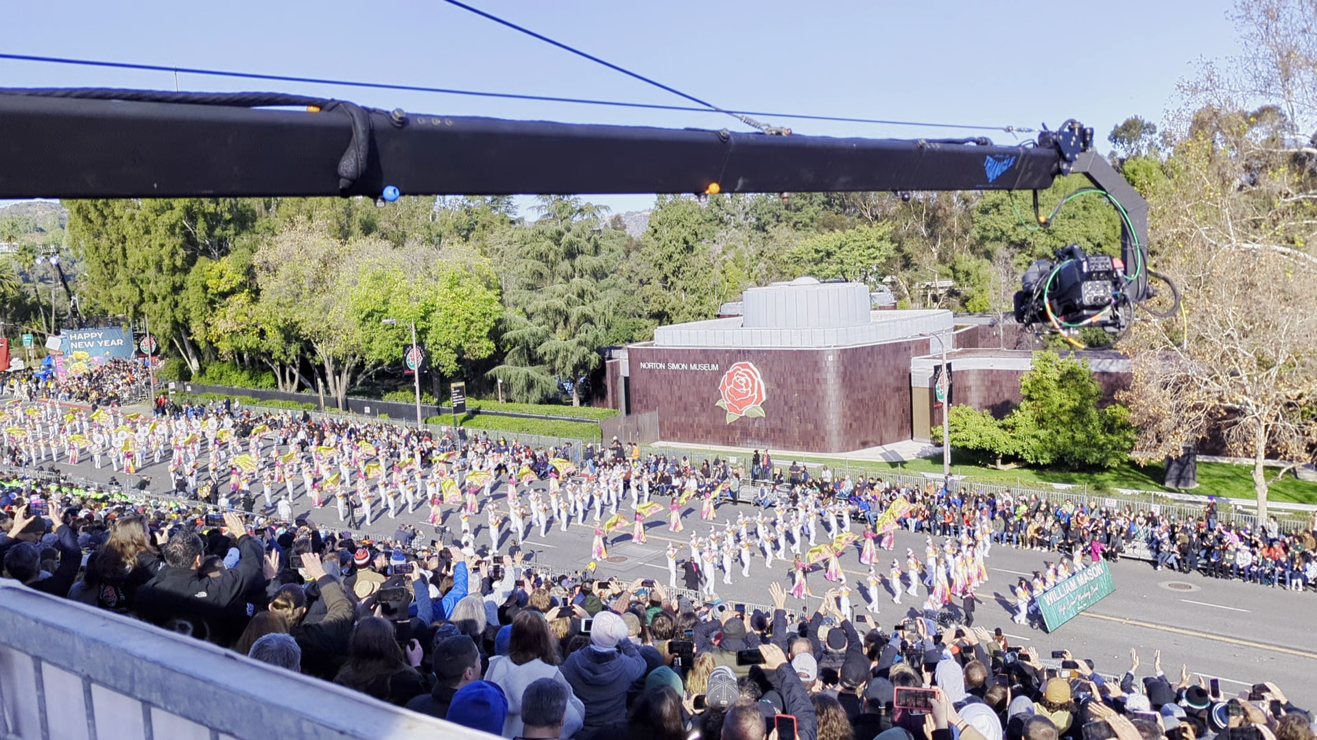 Television camera crane extends over a marching band and packed grandstands during the 2024 Rose Parade in Pasadena.