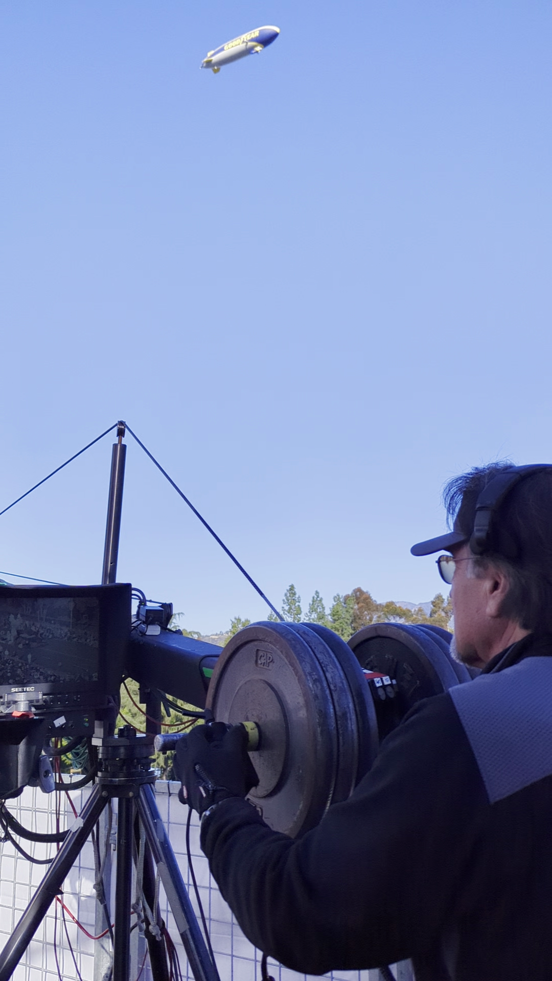 Close-up of a camera crane crew member working with equipment while the Goodyear blimp flies overhead during the 2024 Rose Parade.
