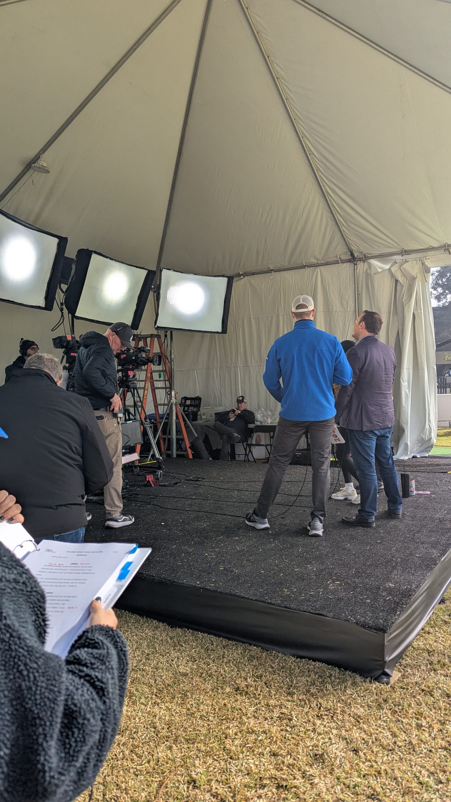 Behind-the-scenes view of a KTLA Rose Parade television production setup beneath a large white tent, featuring studio lights, broadcast cameras, crew members and a production script on a clipboard in the foreground.