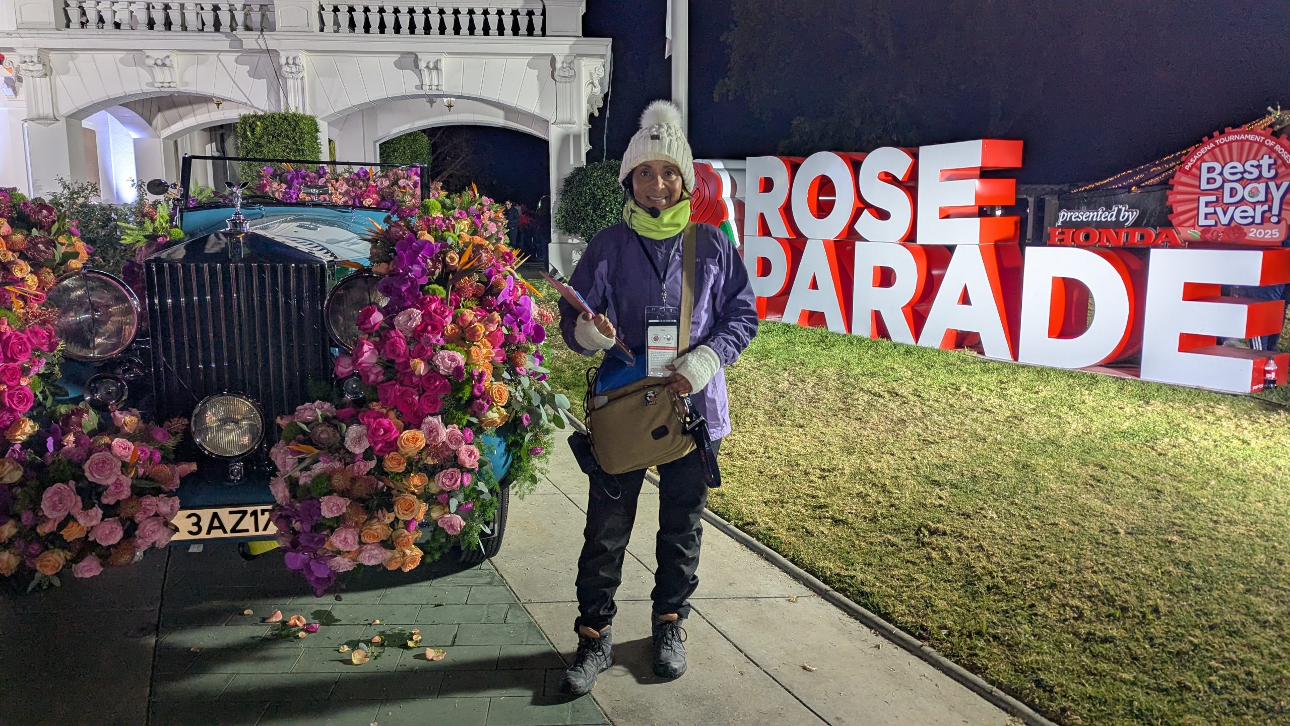 Patricia Nazario wearing production credentials and a headset beside a flower-covered vintage Rolls-Royce and illuminated Rose Parade sign.