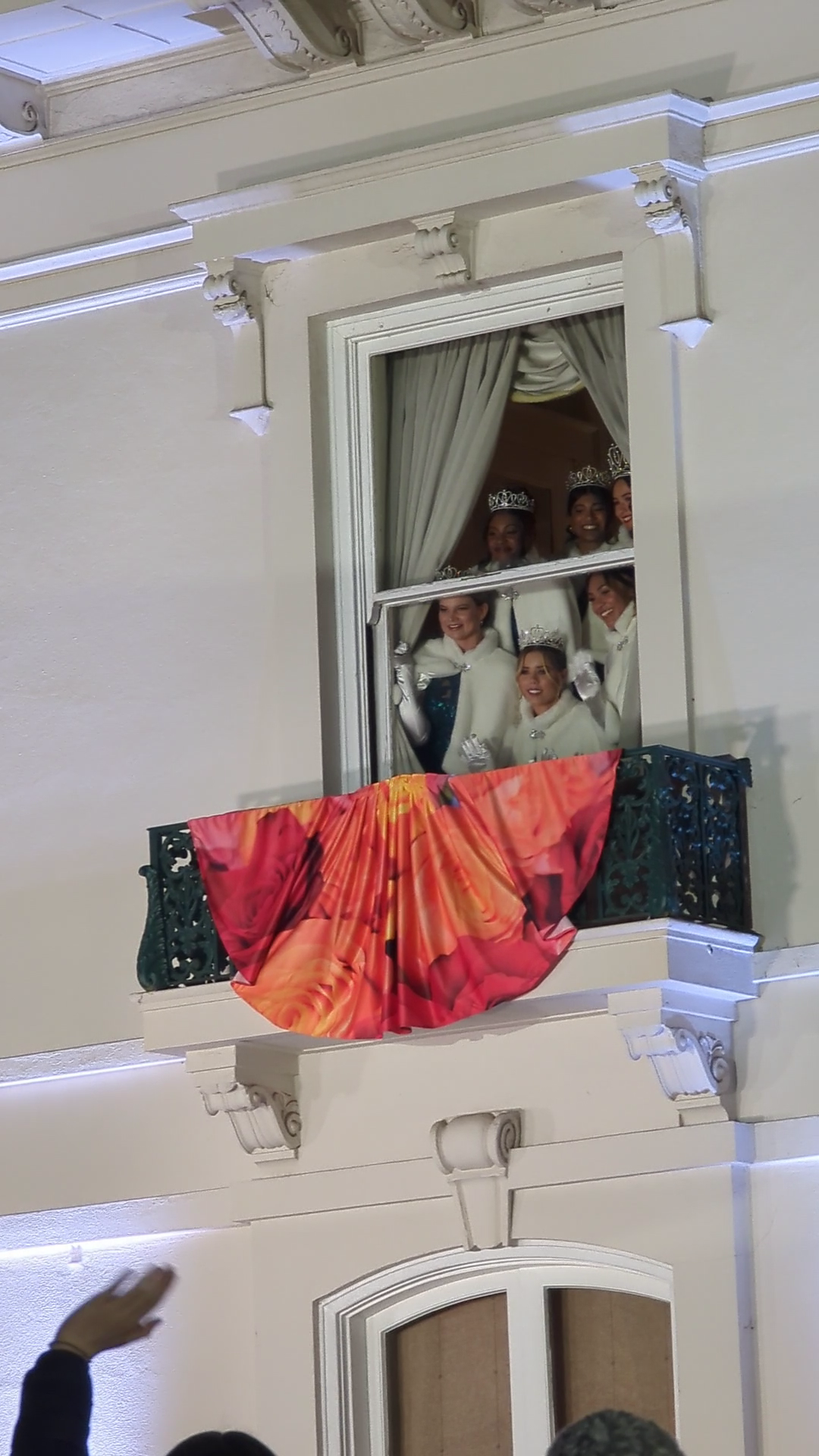 The 2025 Rose Queen and Royal Court, wearing tiaras and white coats, wave to spectators from a decorated balcony window at Tournament House in Pasadena.