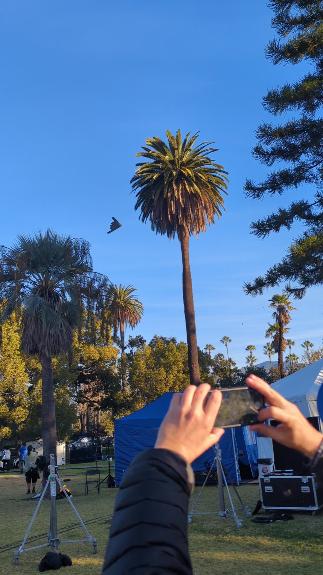 B-2 stealth bomber flying above palm trees in Pasadena as a crewmember captures the aircraft with his cell phone just feet away from the television production tents.
