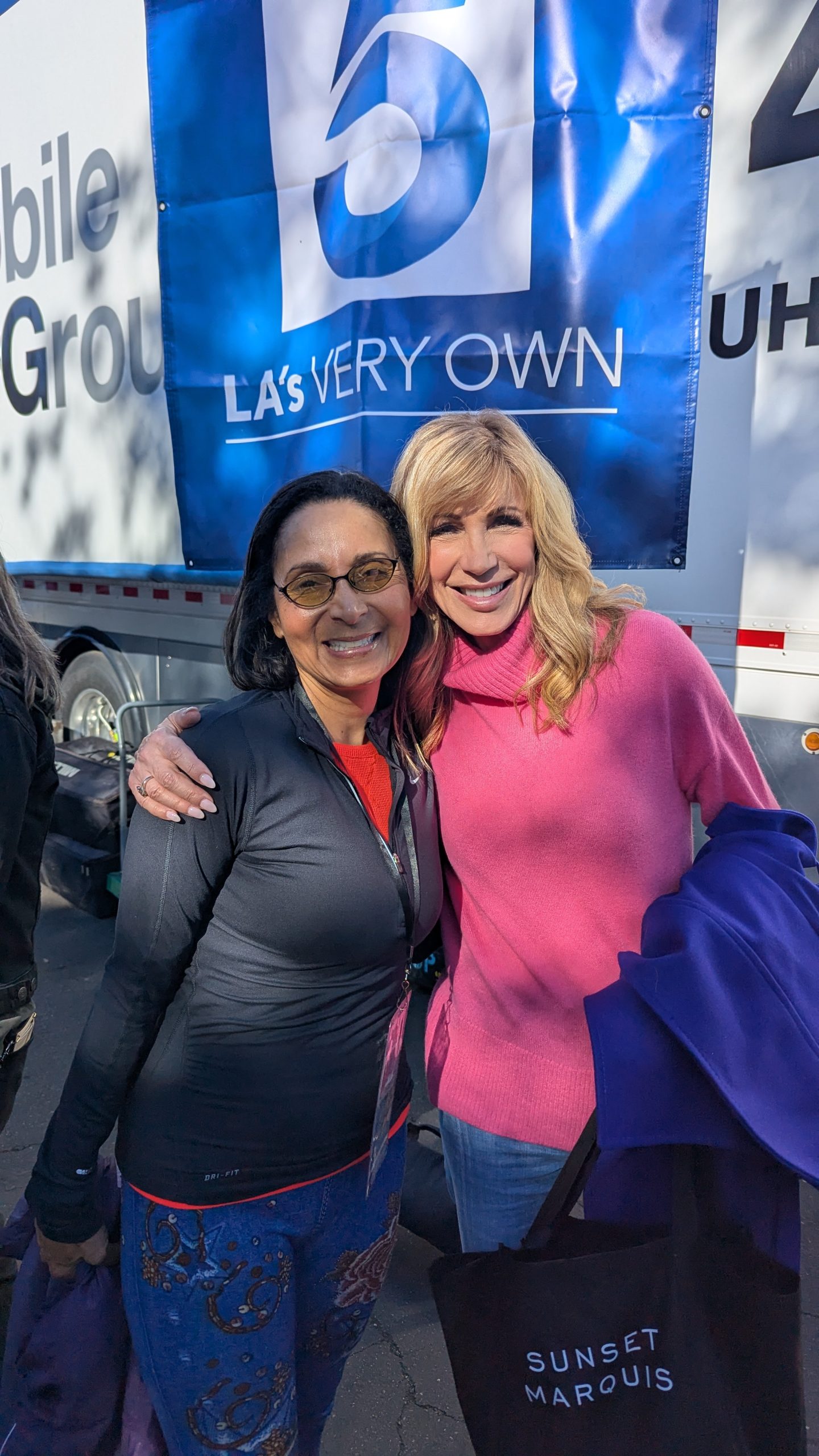 KTLA field producer Patricia Nazario, dressed in black, poses with television personality Leeza Gibbons in a pink sweater in front of a KTLA 5 broadcast banner after the 2025 Rose Parade production.