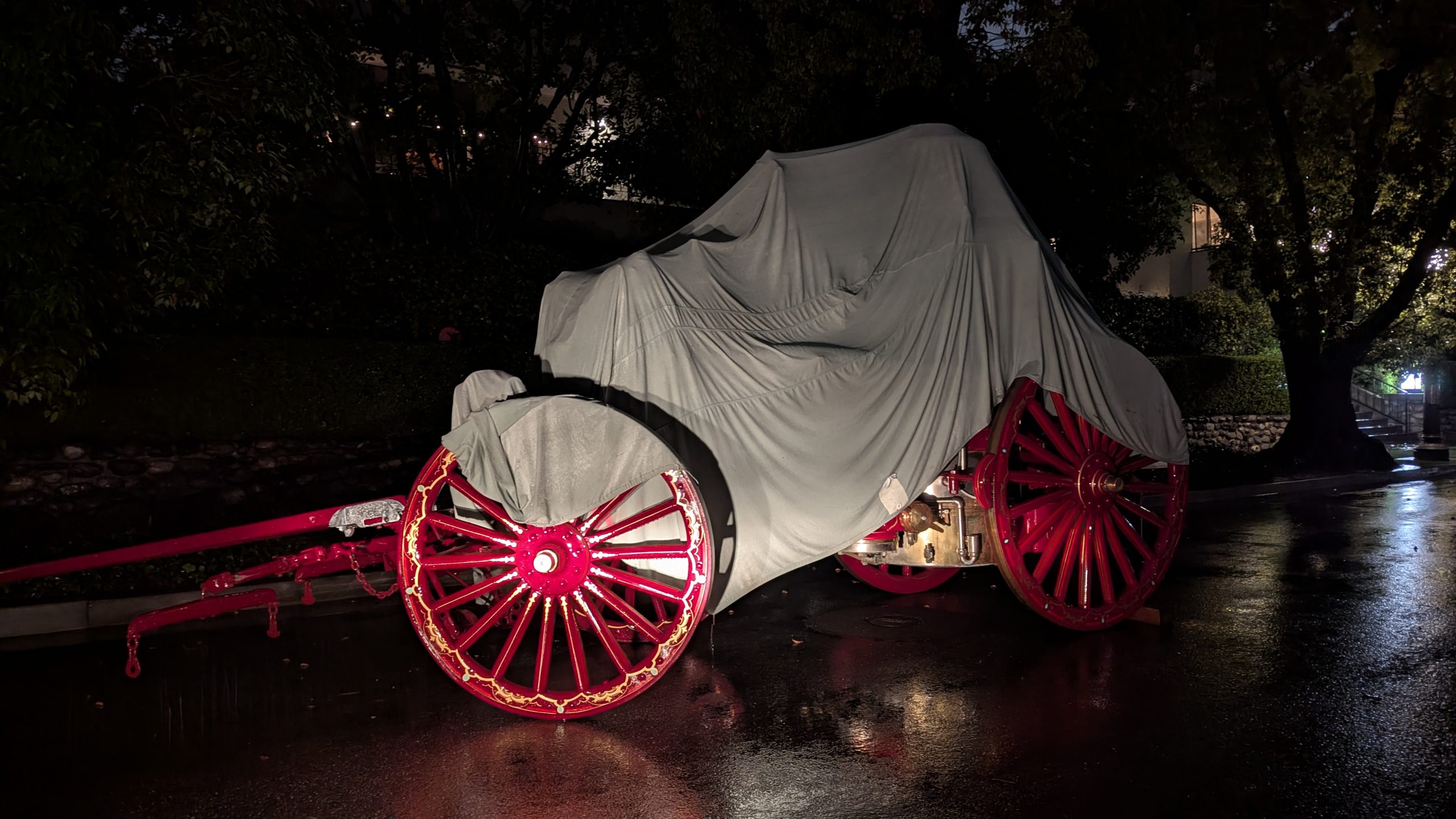 Covered carriage with red wheels on wet pavement during predawn preparations for the 2026 Rose Parade in Pasadena.