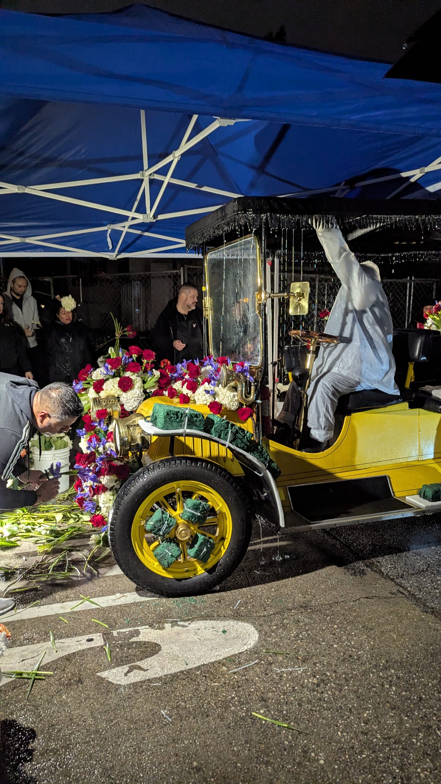 Yellow vintage car decorated with flowers beneath a blue canopy during rainy predawn preparations for the 2026 Rose Parade.