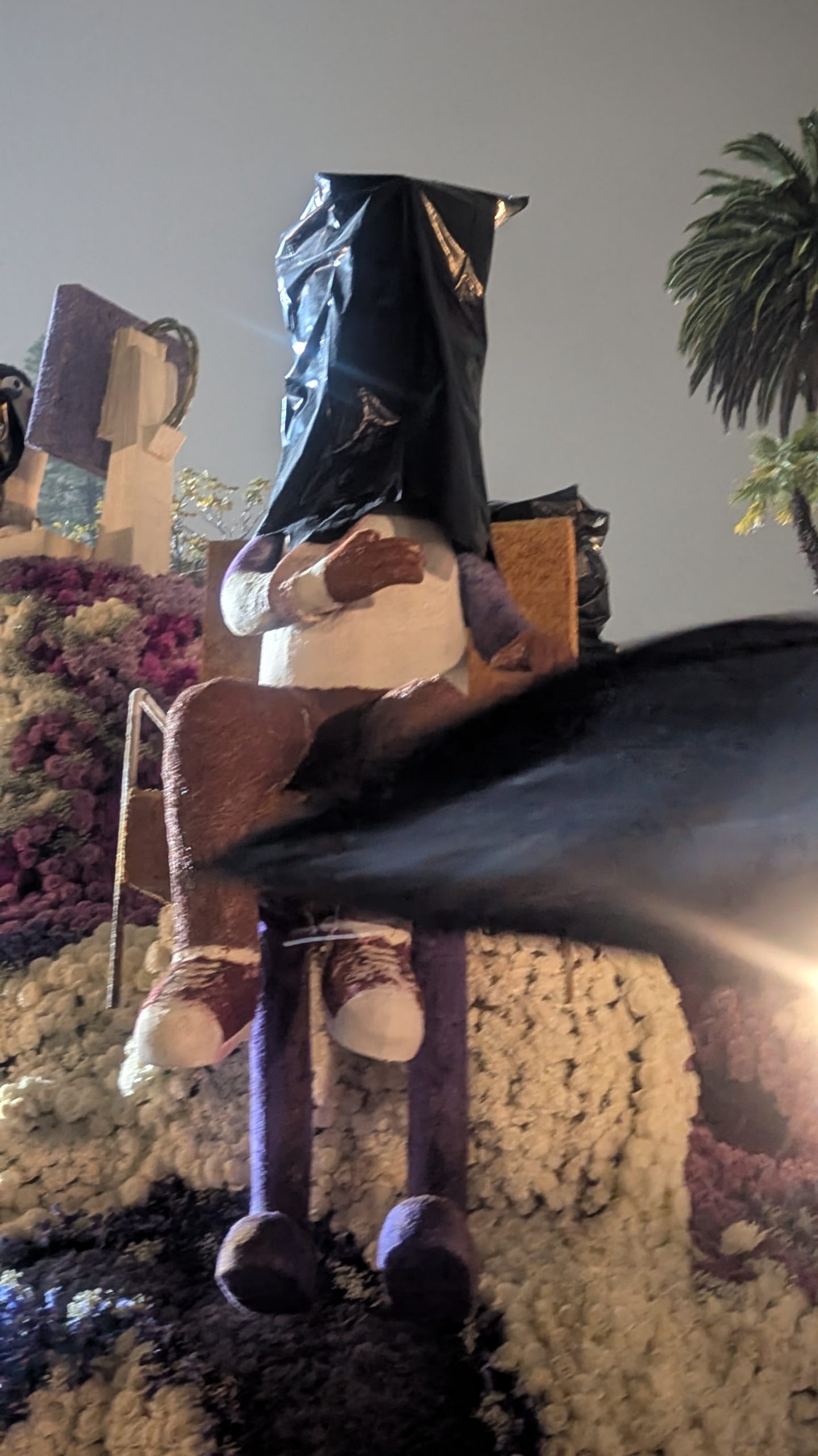 A seated figure on a flower-covered Rose Parade float has its head protected by a black plastic covering during rainy conditions ahead of the 2026 parade in Pasadena.